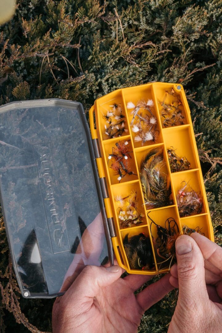 A hand holding a yellow fishing tackle box with various fishing lures and bait.