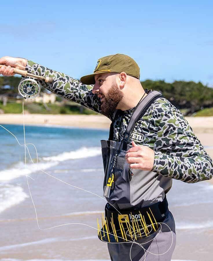 Man wearing fishing gear casting a net by the shore on a sunny day.